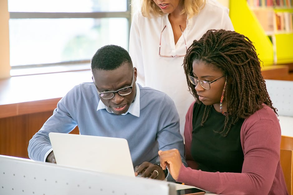Crop unrecognizable female tutor in white shirt helping young African American students in eyeglasses doing research using laptop in light classroom